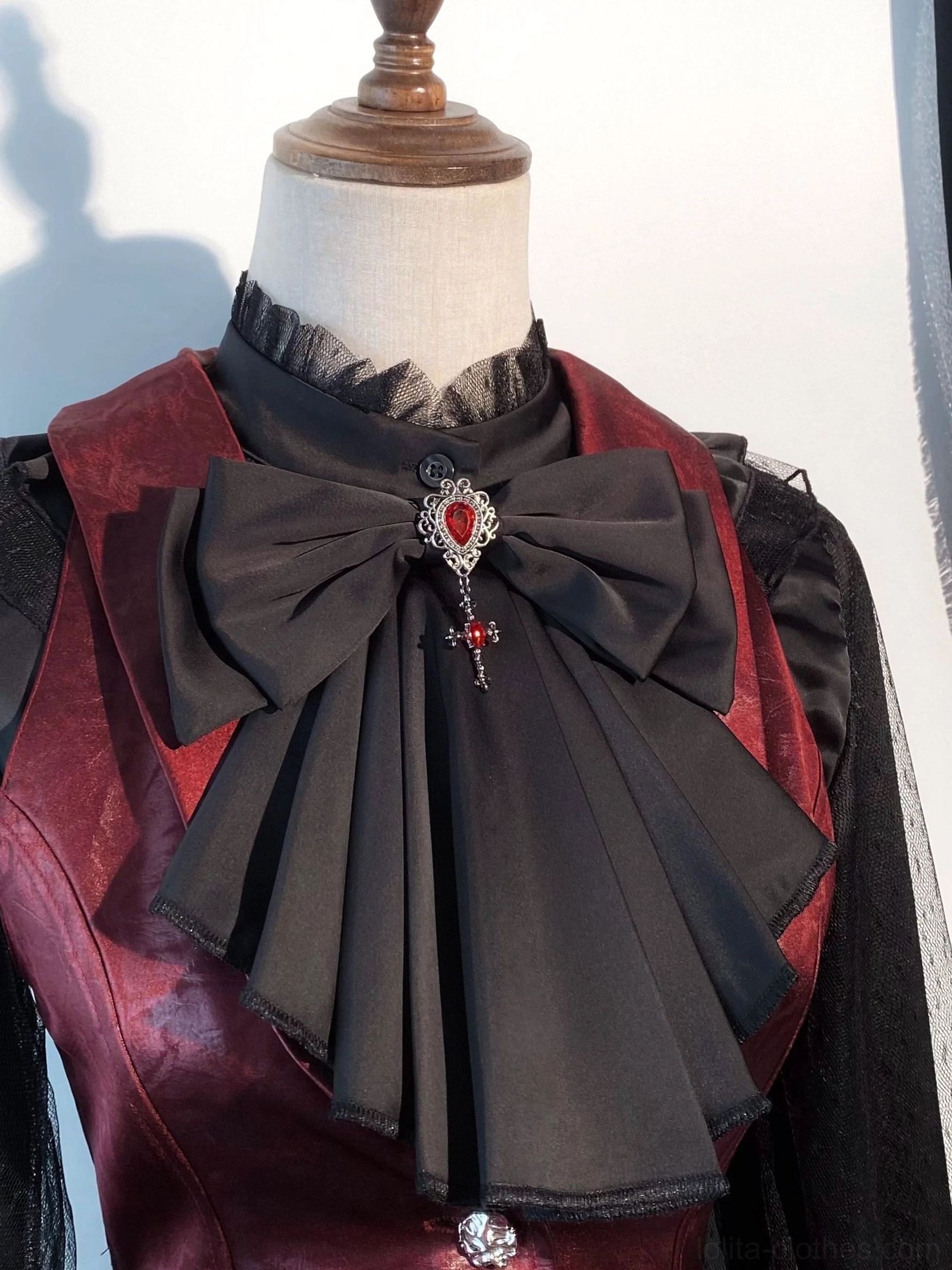 Close-up front view of wine red leather gothic lolita waistcoat with ruffled high stand collar, black bow with red gemstone brooch accent, metal button details