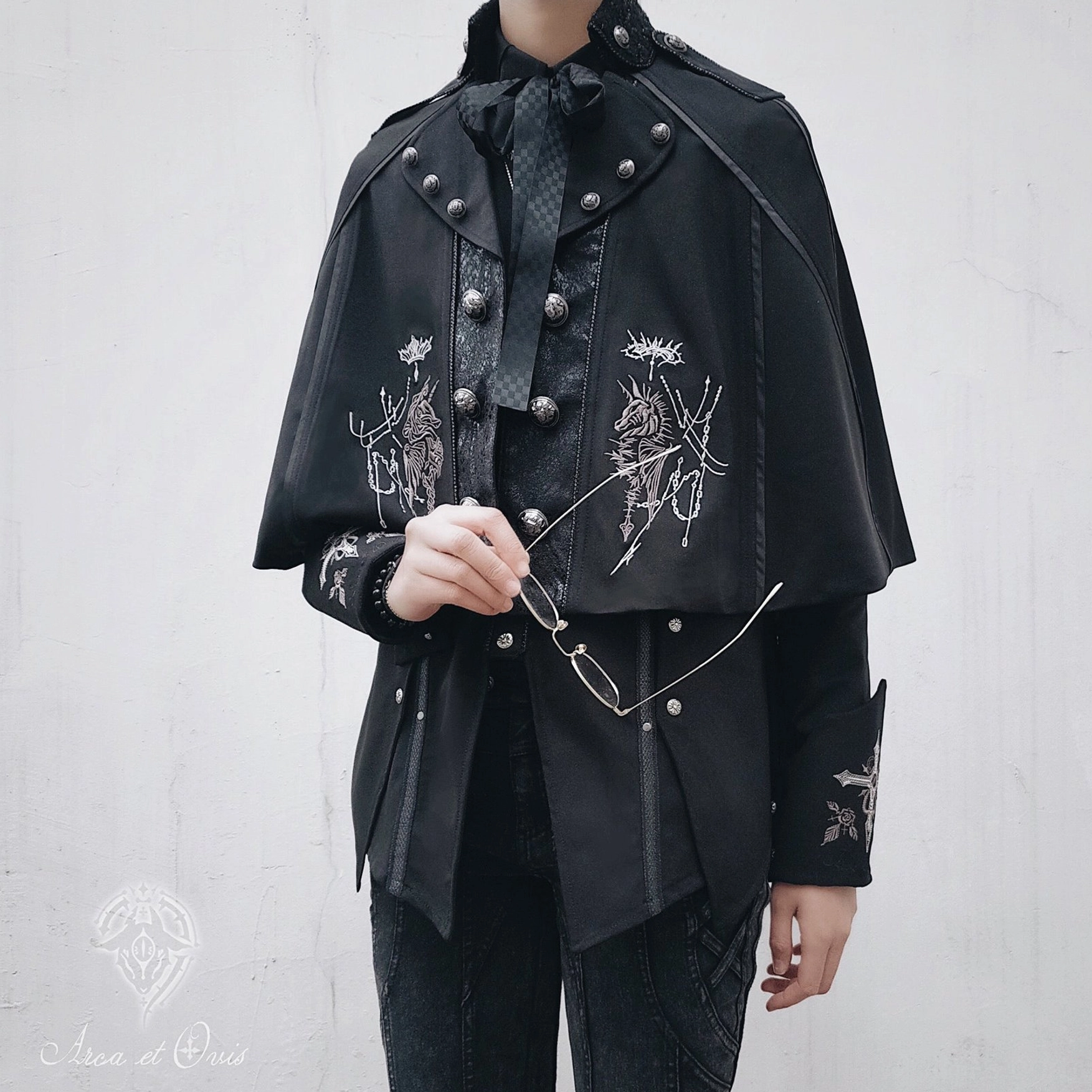 Gothic Ouji Lolita short black embroidered cape with silver knight motif front half-body view on androgynous male model, metal stud shoulder decorations and button detailing