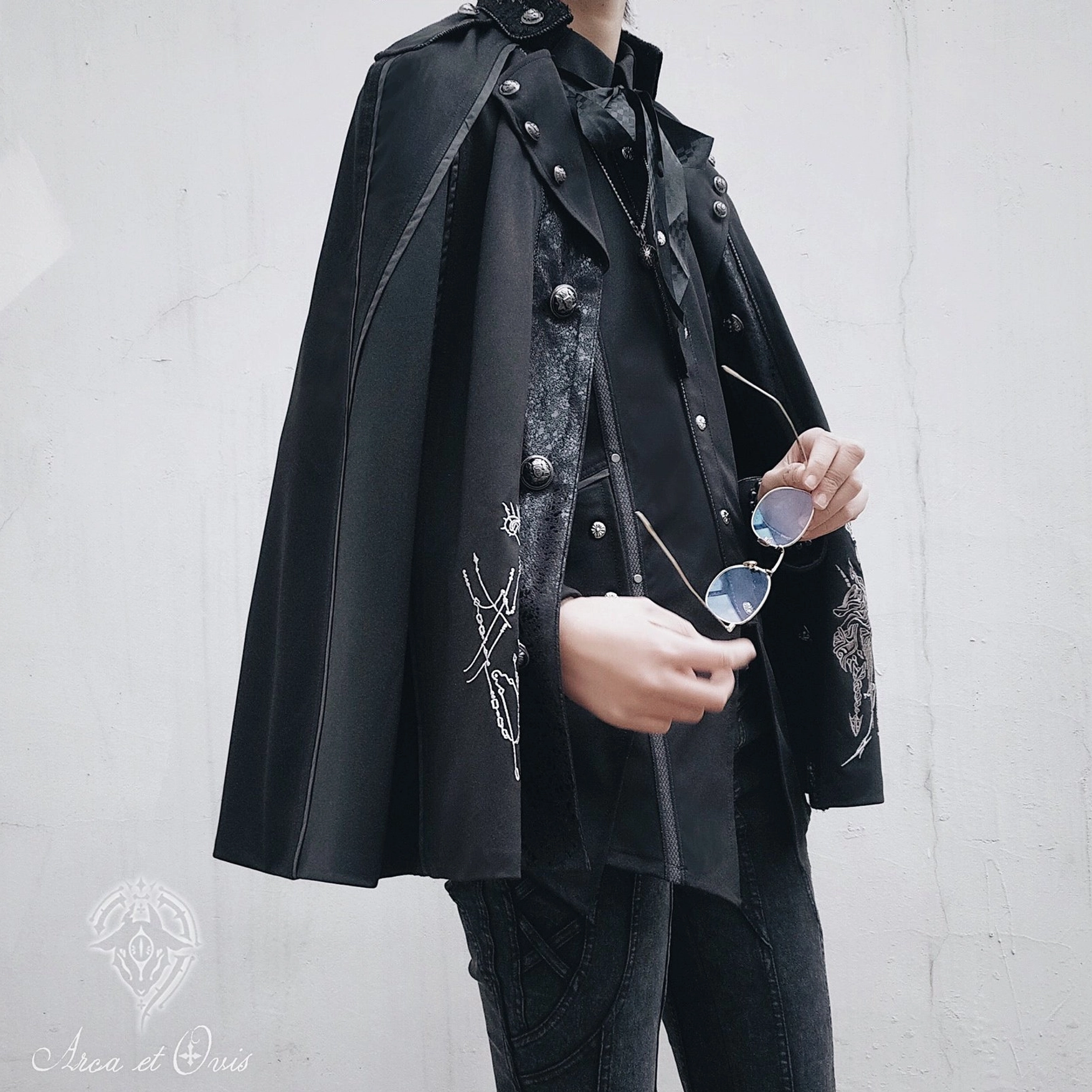 Side half-body shot of black Gothic Ouji Lolita cape with matte fabric, silver knight and floral embroidery, metal button details, layered cape and inner shirt set on model with sunglasses