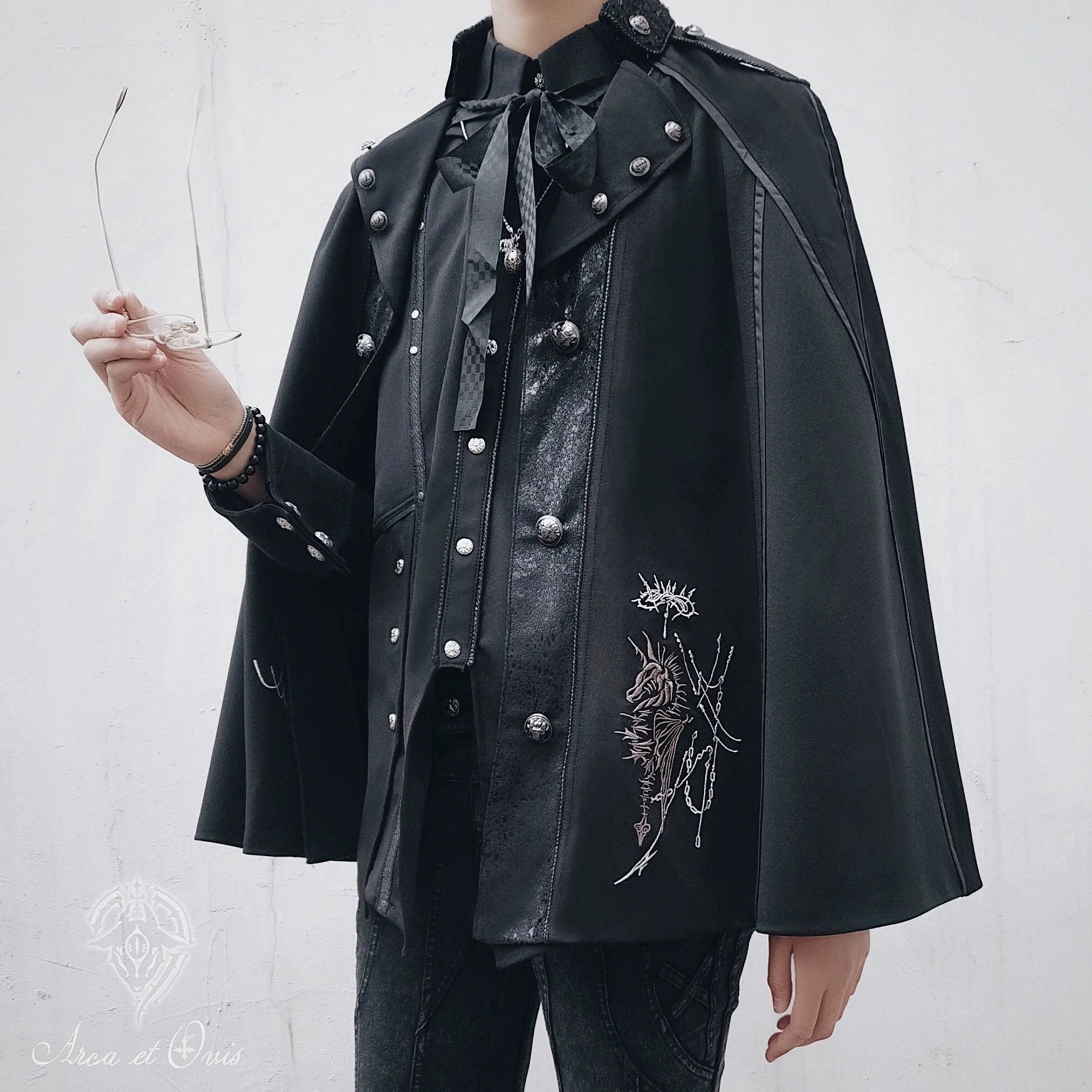 Gothic Ouji Lolita long version black embroidered cape front half-body on androgynous male model, silver knight and floral embroidery, metal stud shoulder decorations, matching bow necktie
