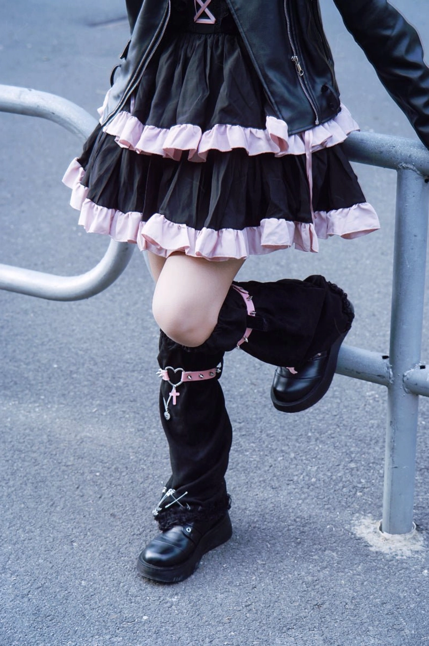 Close-up low angle shot of gothic leg warmers with lifted leg showing pink cross charms ribbon lace-up details suede texture and metal accents outdoor guardrail