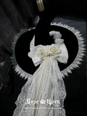 Flat lay display of black and white gothic lolita witch hat headdress showing wide scalloped lace-edged brim, double white roses, lace bow and embroidered lace veil trailing detail from top-down view