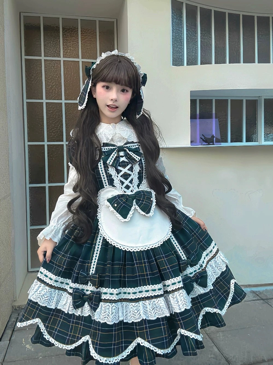 Dark green plaid maid-style lolita OP dress with twin hairstyles, white lace trim, black bow accents, scalloped petal hem, and apron front on model