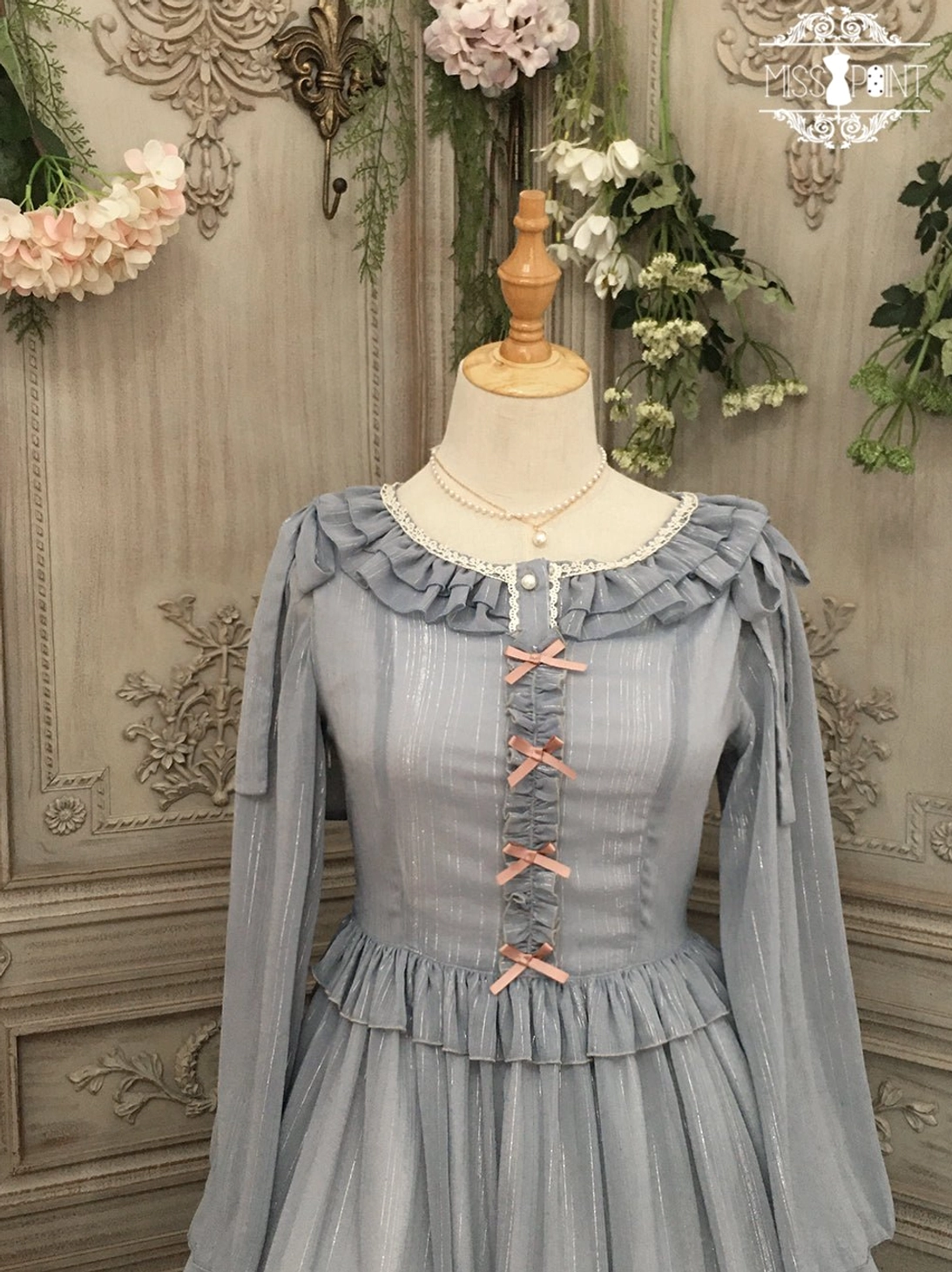 Grey lolita cardigan with ruffled neckline and shoulders, featuring pink ribbon lace-up front detail and ruffled waist trim, shown as part of complete lolita coordinate set, displayed on mannequin with vintage floral scene backdrop