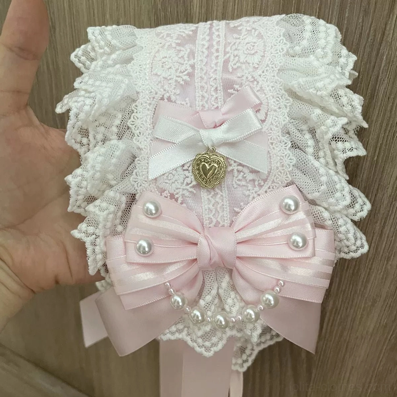 Handmade Sweet Lolita lace hairband held in hand, ruffled white lace structure, double satin bows in pink and white, pearl trim accents, gold heart pendant and adjustable pink ribbon straps