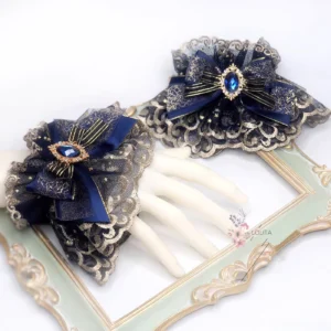 Pair of indigo gothic lolita wrist cuffs with gold-embroidered scalloped lace, navy satin bows, and teardrop blue gem accents displayed on white mannequin hands with vintage gilt frame styling