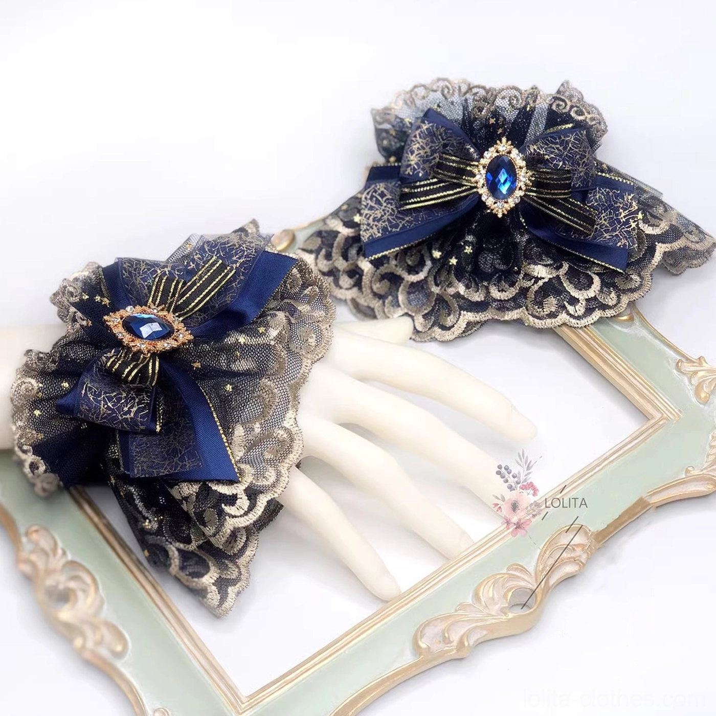 Pair of indigo gothic lolita wrist cuffs with gold-embroidered scalloped lace, navy satin bows, and teardrop blue gem accents displayed on white mannequin hands with vintage gilt frame styling