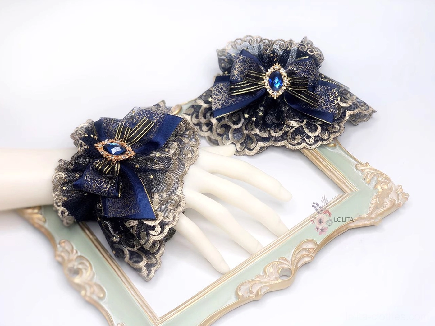 Pair of indigo gothic lolita wrist cuffs showcasing gold-embroidered mesh lace, navy satin bows with teardrop blue gems, and scalloped detailing on partial white mannequin hand