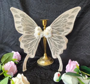 Cream ivory embroidered butterfly hair accessory with delicate sheer lace wings and white fabric flower ornaments displayed frontal view against dark velvet backdrop with gold candlestick