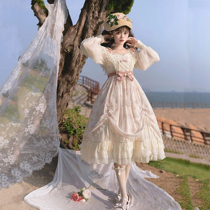 Ivory floral princess lolita OP dress front three-quarter view with cream base color, pink waist bow, ruffled square neckline, sheer puff sleeves, outdoor seaside setting with lace backdrop and model wearing floral straw hat white stockings