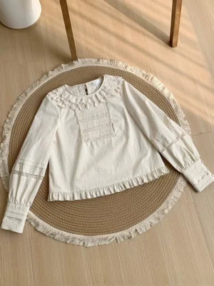 Ivory lolita cotton blouse flat lay top-down view showing cropped silhouette with ruffled lace collar trim and detailed sleeve cuffs on woven rug
