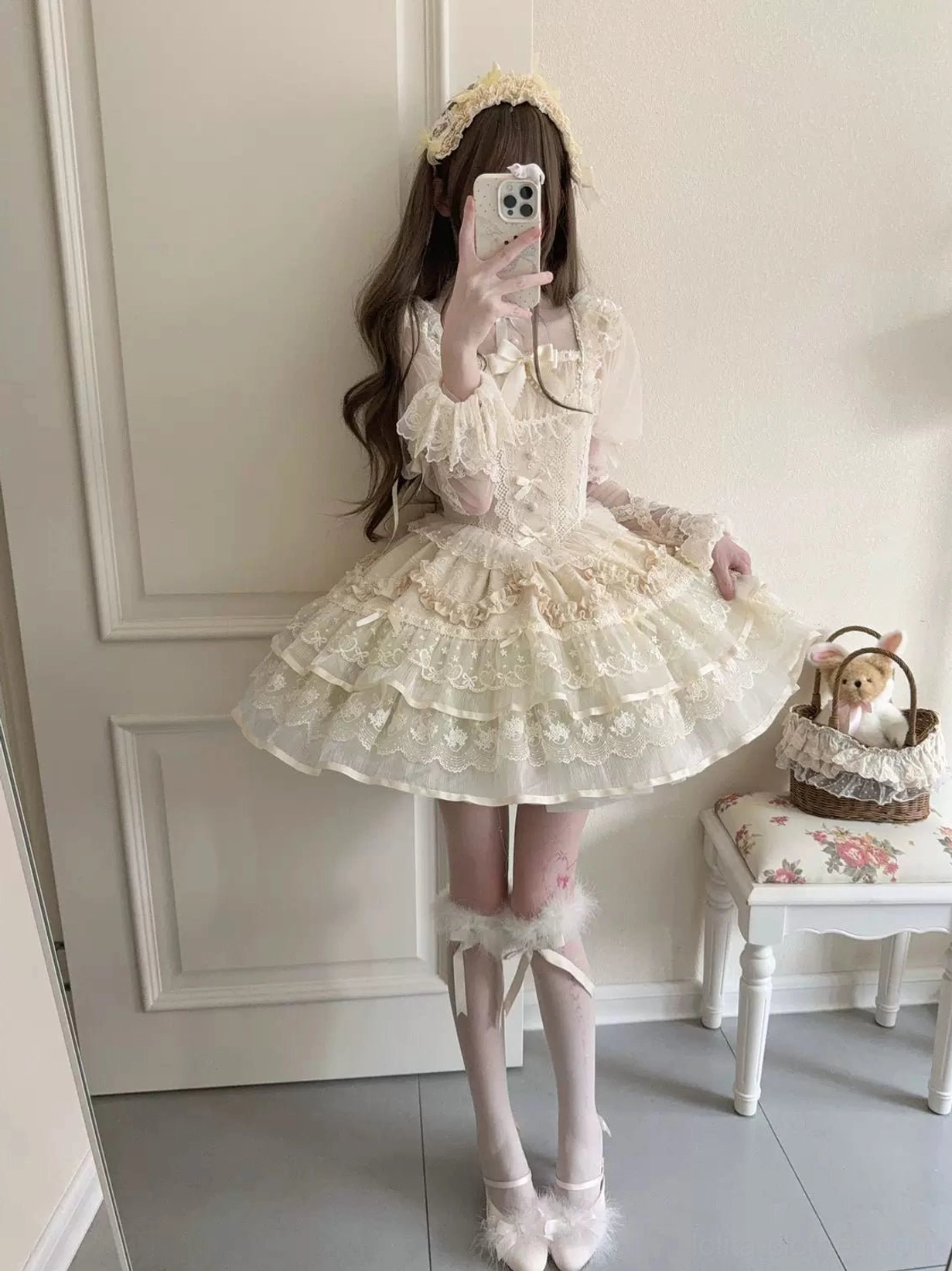 Front mirror selfie of female model in ivory sweet lolita JSK dress with golden hair accessory, multi-layered ruffled lace petal skirt, fluffy leg warmers, ballet flats, and vintage-inspired styling