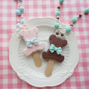Overhead view of dual kawaii bear biscuit ice cream resin necklaces in pink and chocolate brown with mint blue bows on white embossed plate and pink gingham tabletop displaying handmade lolita pendant necklace set