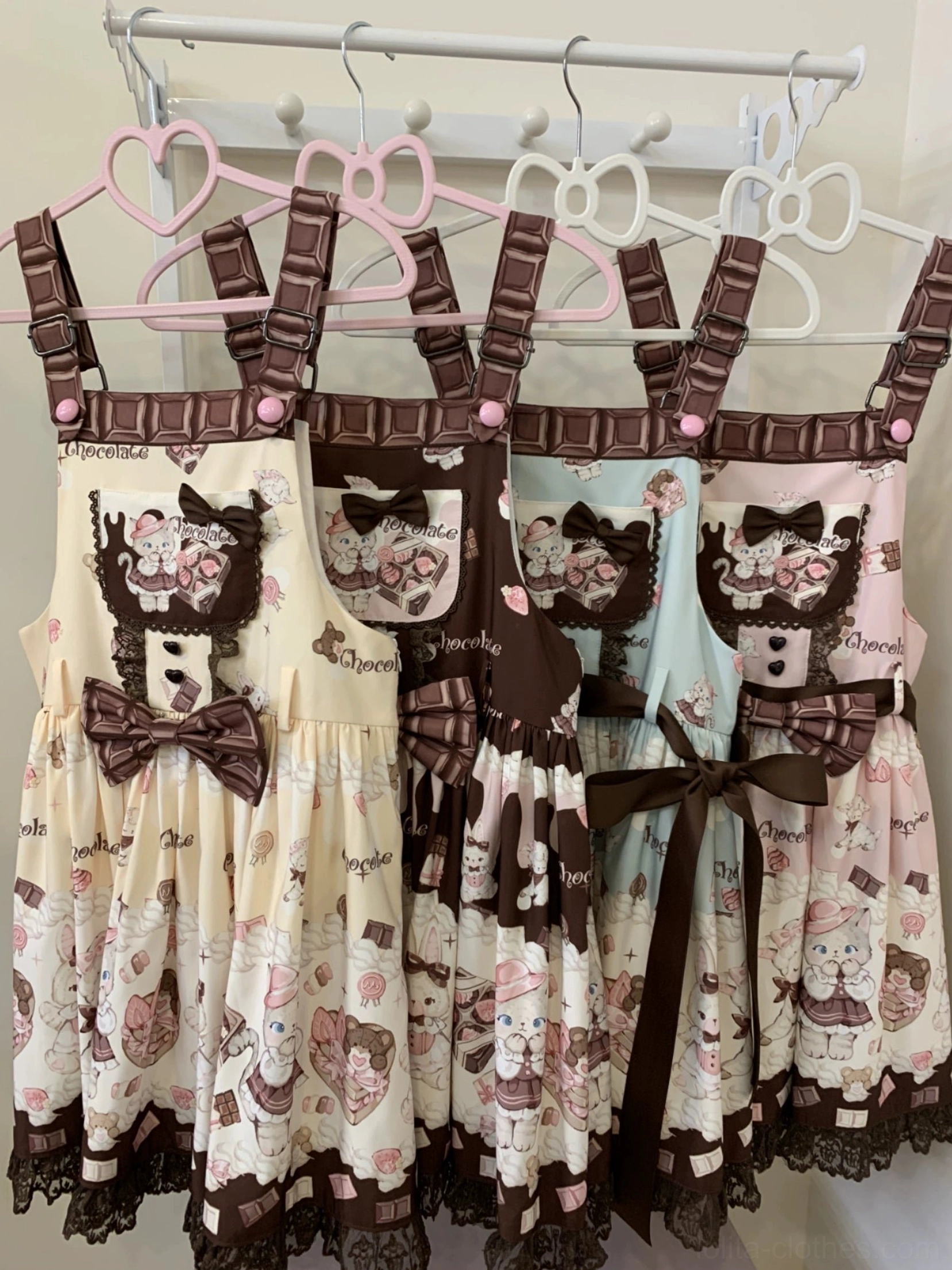 Multiple colorways of cute cat and dessert printed Lolita salopette dresses displayed on clothing rack, showing chocolate brown shoulder straps, lace trim details, and detachable bow accents in cream yellow, mint green, pink, and coffee brown colors