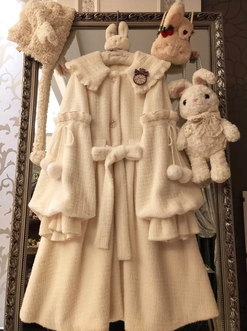 Full front hanging display of cream fluffy lolita winter coat with bow knot waist decoration, pom-pom pendants, layered fluffy hem, and detachable beret against vintage mirror
