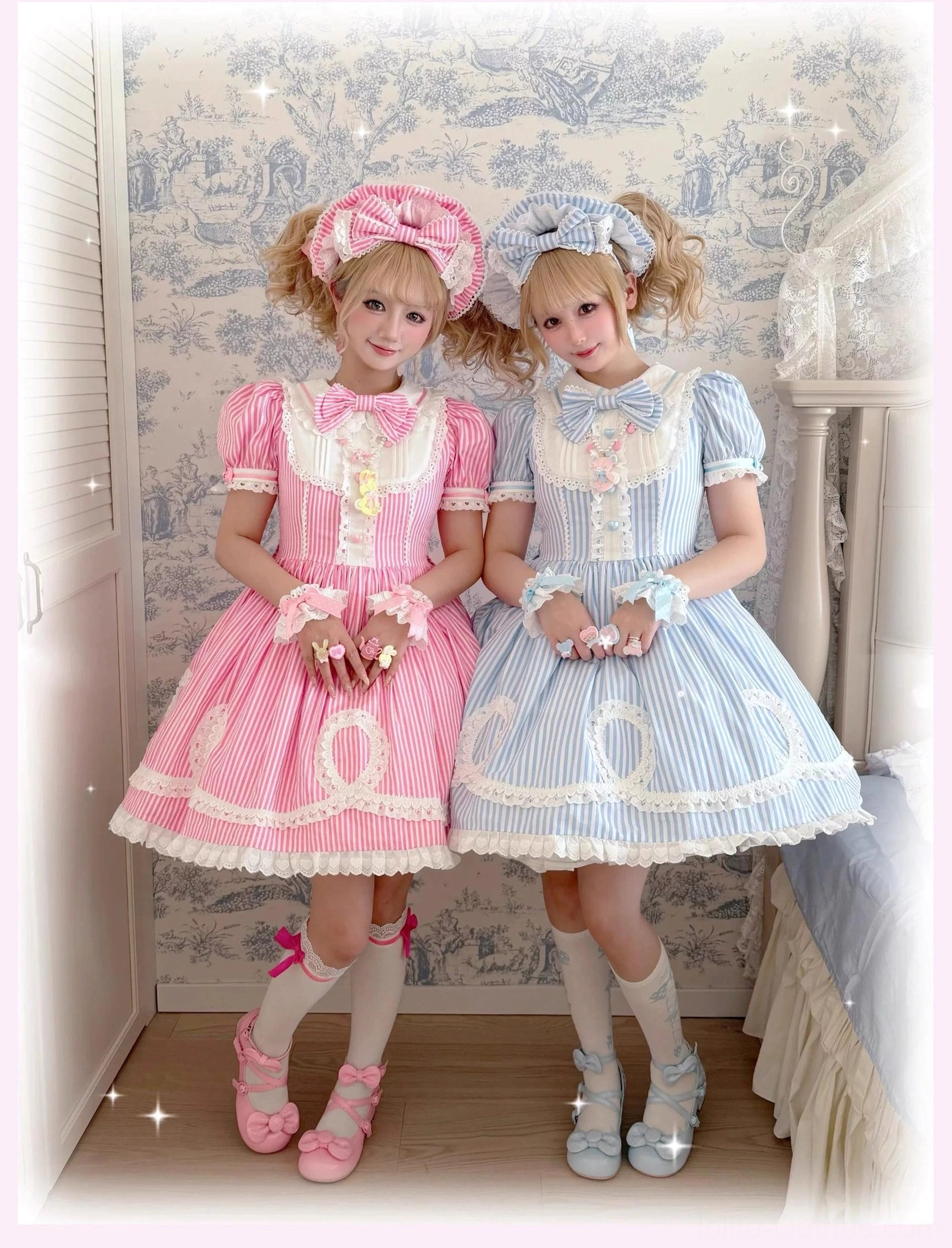 Two kneeling female models wearing pink and blue striped old school lolita OP dresses with matching bonnet accessories and heart-shaped button cuffs