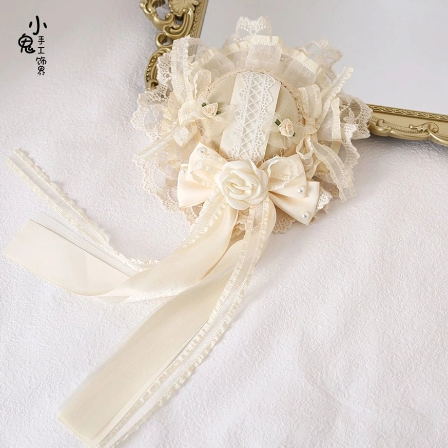 Lace rose headdress ornament with ruffled lace border, satin bow, fabric rose and flowing ribbon tails displayed on white background with gold mirror frame