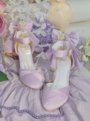 Lavender classic Lolita wine glass heeled sandals flat lay display on purple fabric with flower arrangements and pearl chains, showing detachable bow with rhinestone ornament and elegant heel silhouette