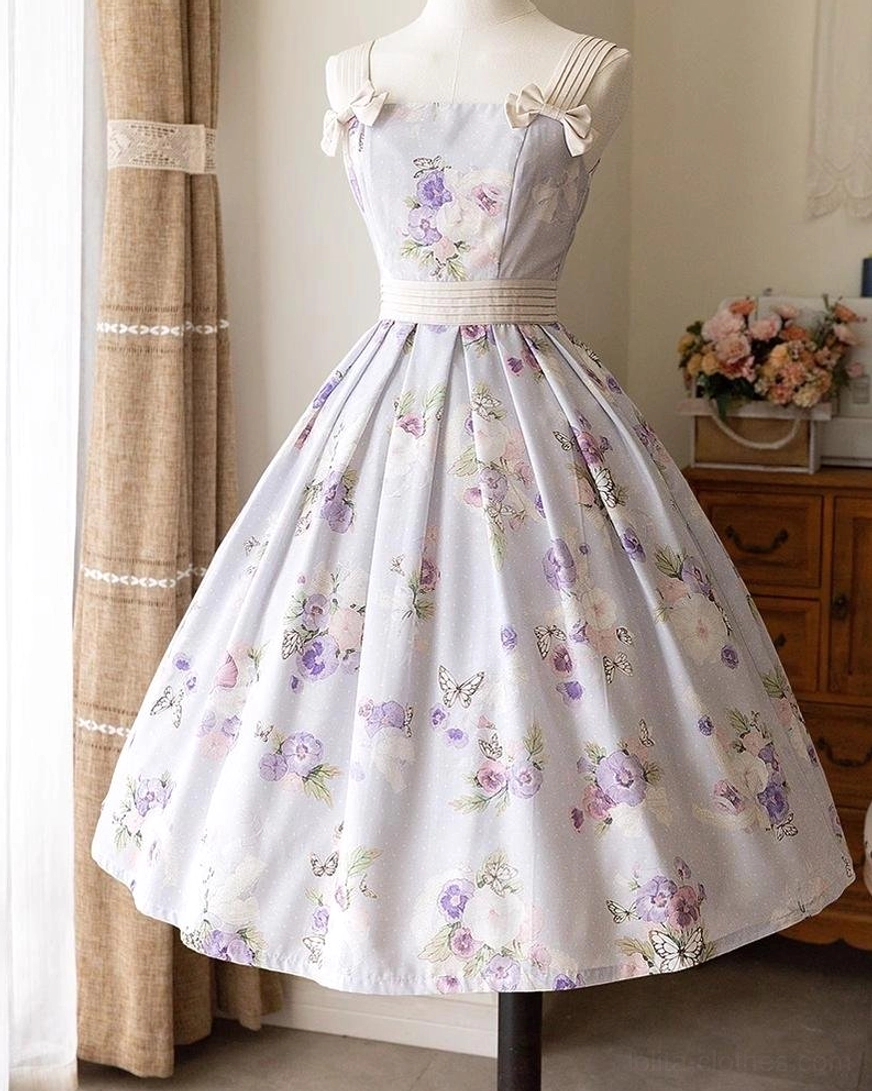 Lavender floral lolita jumper skirt with romantic purple flower prints, square neckline with bow-adorned straps, matching waist belt, and flowing flared mid-length skirt captured in full front view