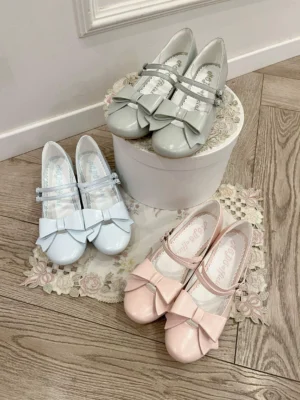 Three sets of Lolita Mary Jane shoes in light blue light pink light gray with low block heel and decorative bow strap displayed on wooden floor with lace doily and bucket props