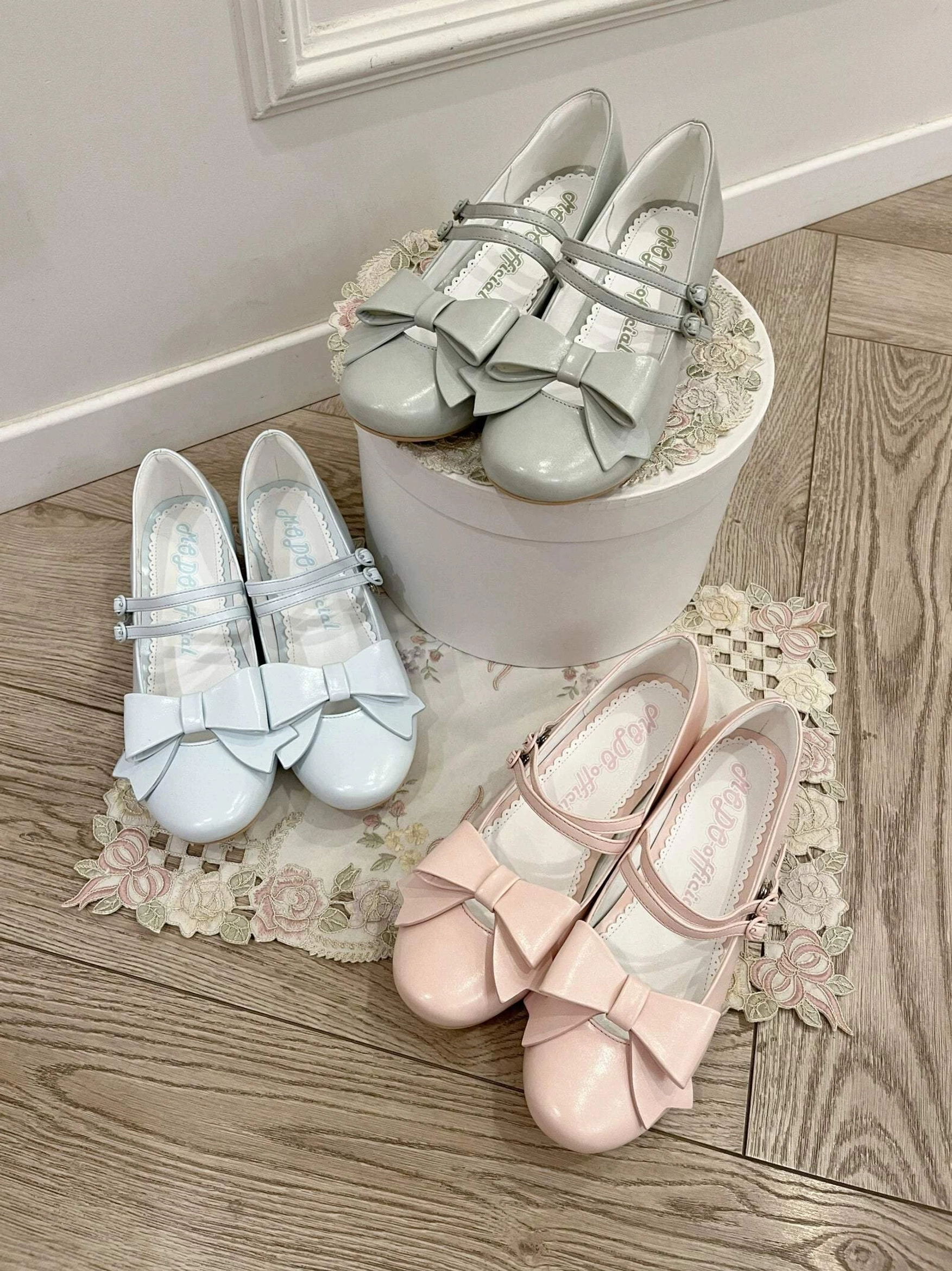 Three sets of Lolita Mary Jane shoes in light blue light pink light gray with low block heel and decorative bow strap displayed on wooden floor with lace doily and bucket props