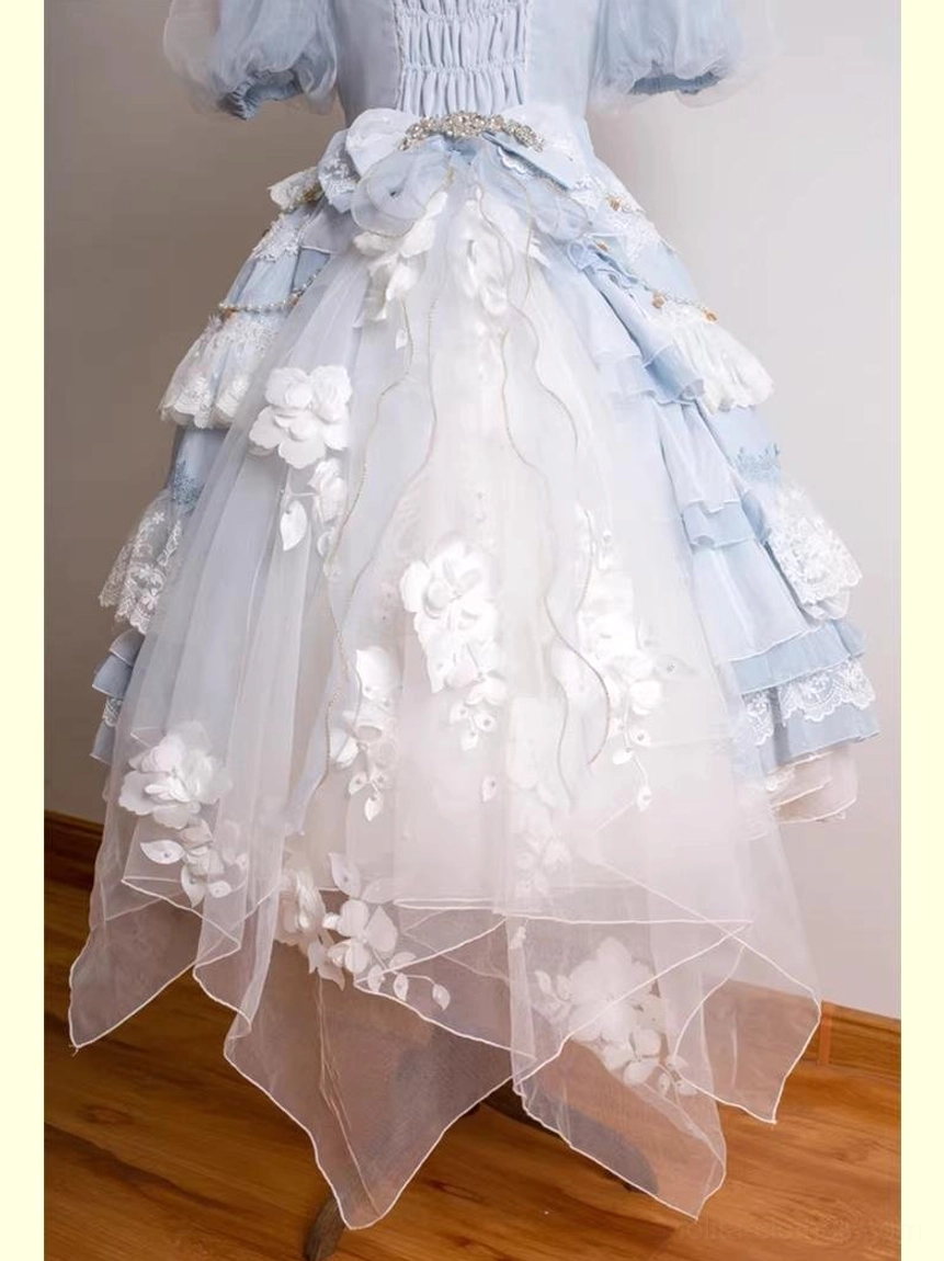 Light blue classic lolita dress lower part front close-up showing asymmetrical sheer mesh train with white 3D floral appliqués, layered ruffled skirt, and rhinestone bow brooch at waist
