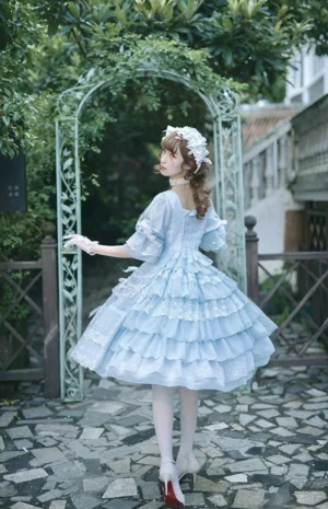 Light blue Sweet Lolita OP dress with multi-layered ruffle skirt and white stockings with red heels, rear three-quarter garden arch view