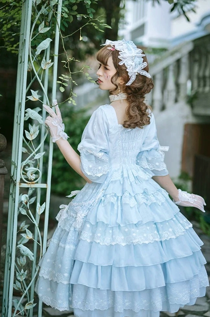 Light blue Sweet Lolita OP dress with tiered layered lace skirt, half puff sleeves, and white lace gloves, rear side view garden fence