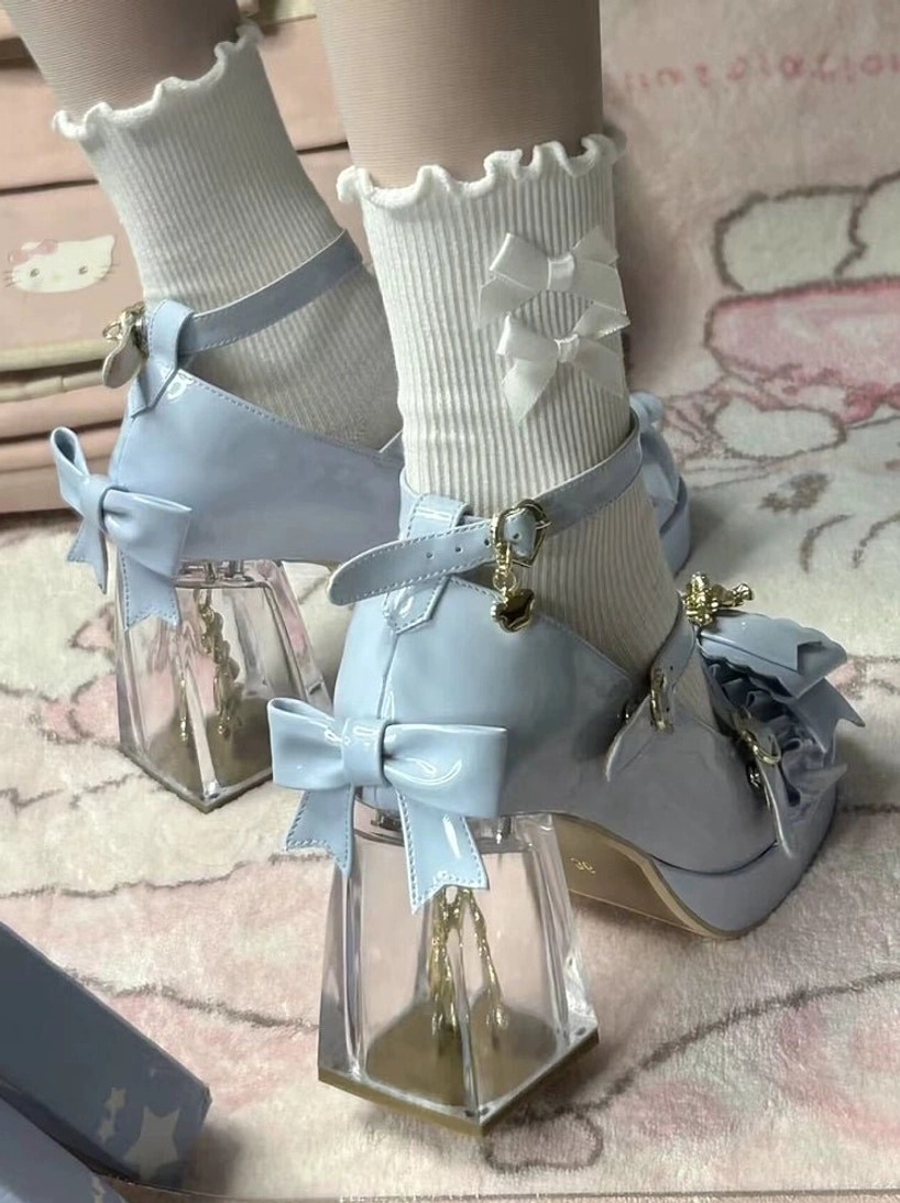 Light blue sweet lolita shoes side close-up view, placed next to white frilled ankle socks with bow decoration, transparent square block heel, gold bear charm, crisscross ankle strap design