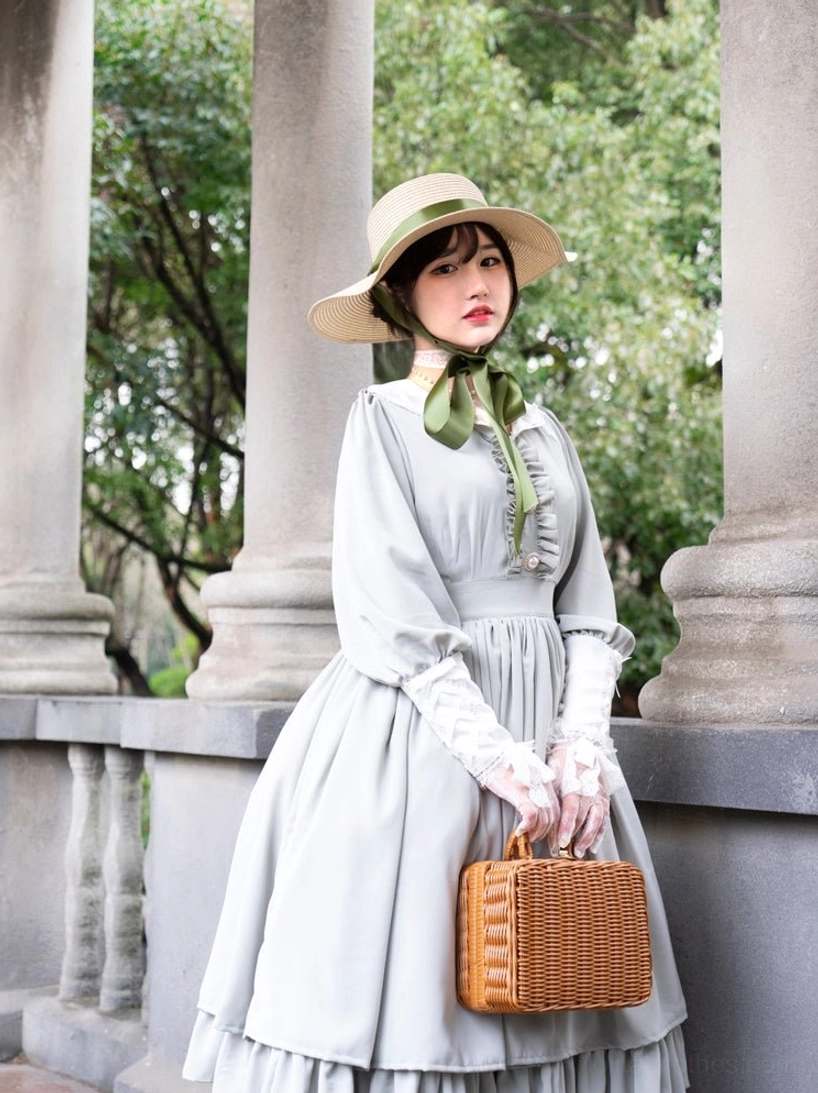 Light gray classic Lolita OP dress half-side front standing view with wide-brimmed straw hat and green neck ribbon, outdoor balcony with wicker bag