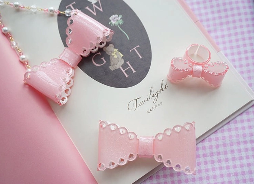 Assorted light pink glittery resin bow necklaces and bow accessories displayed on gingham background with decorative notebook and lolita styling