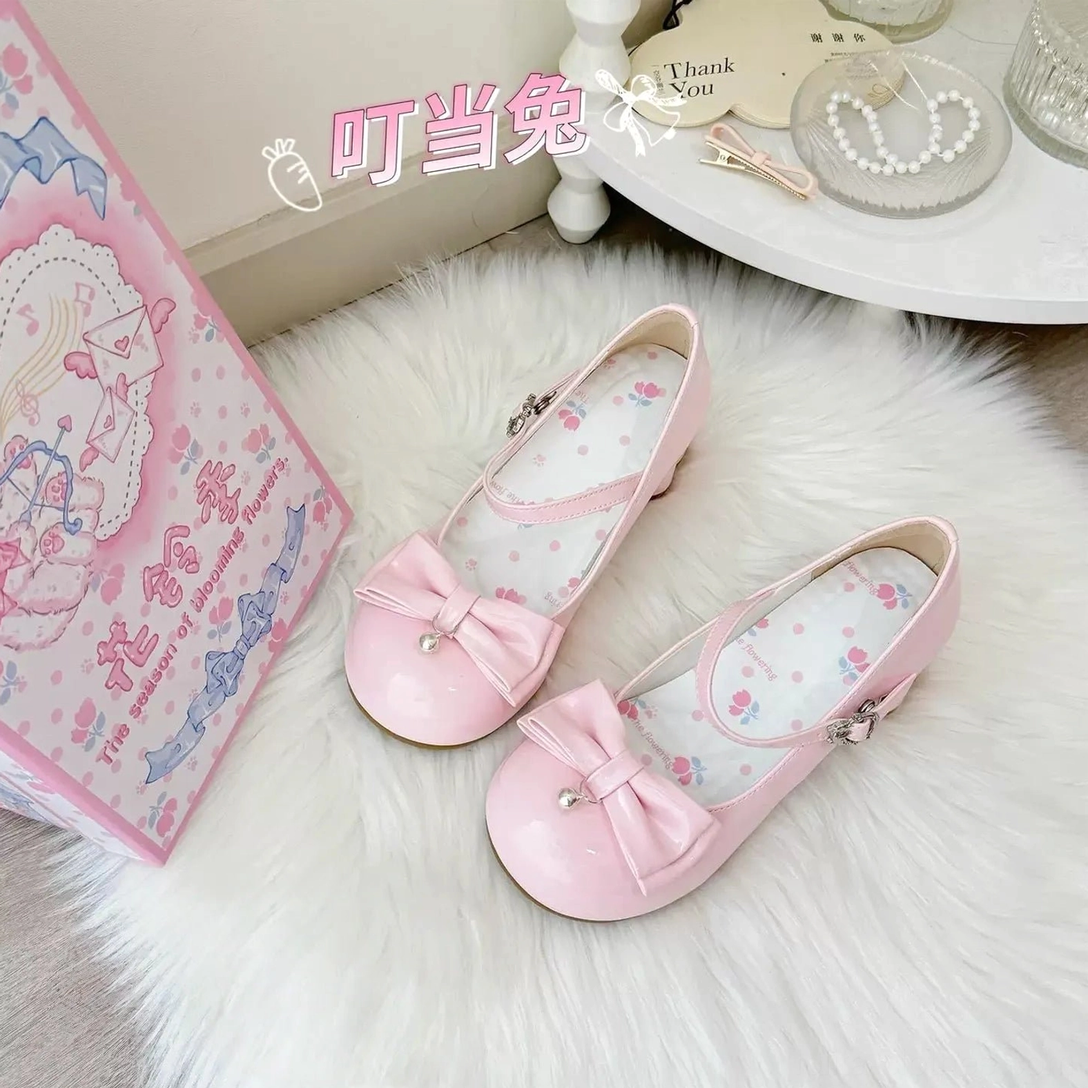Light pink glossy patent leather sweet lolita Mary Jane shoes featuring pearl-accented front bow, printed delicate floral inner lining, ankle strap with buckle closure, chunky low block heel, displayed on white fur rug with themed branded packaging and vintage vanity shelf styling
