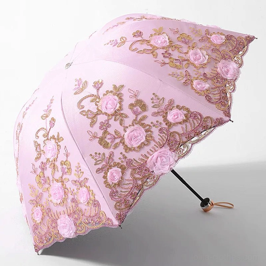 Light pink foldable lolita parasol with gold thread embroidery and three-dimensional pink rose patterns, scalloped lace edge trim, metal handle with hanging strap, fully opened from three-quarter top view against plain white studio background