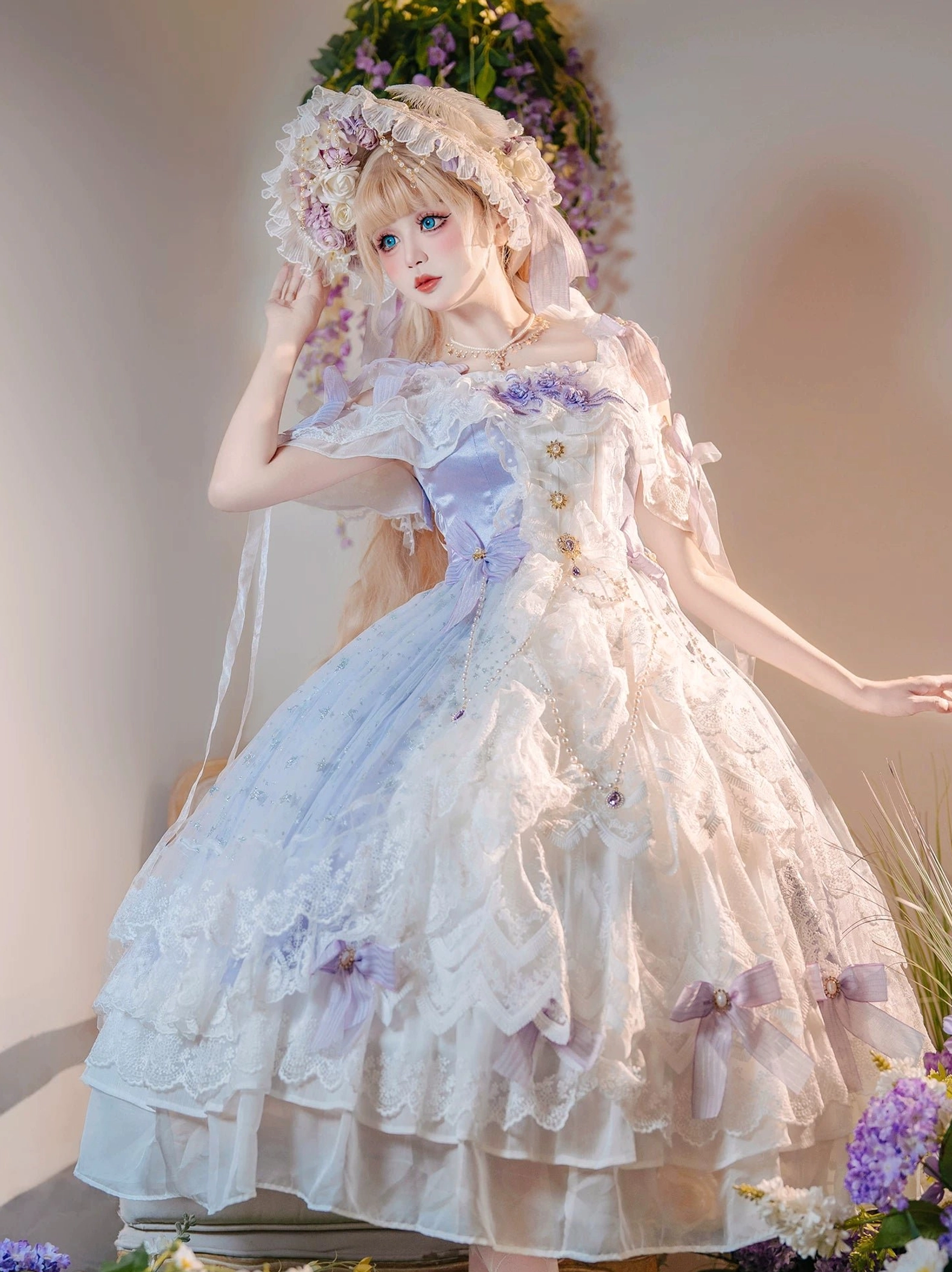 Light purple bridal lolita JSK dress with off-shoulder design, layered lace hem, and purple ribbon bows, displayed on model in studio with floral wisteria backdrop, front three-quarter view