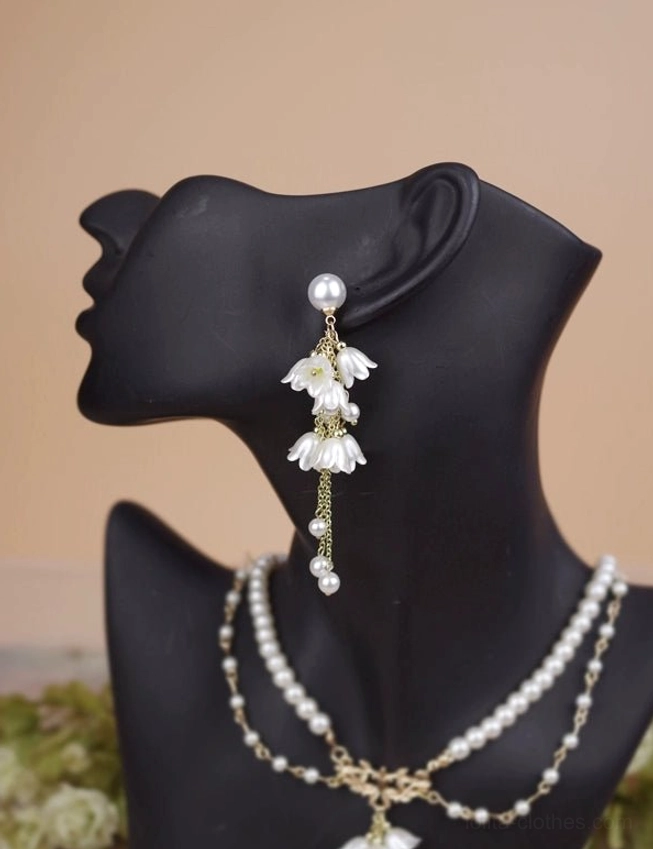 Close-up front side view of lily dangle earrings on mannequin featuring classic pearl stud detailed lily flower pendants delicate gold chain with small hanging pearl accents subtle warm backdrop