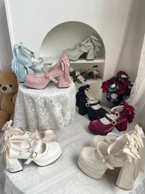 Overhead flat lay display of enchanted lily of the valley classic lolita platform mary jane shoes in multiple color options featuring large detachable bows, gold-tone buckles and thick block heels styled on decorative lace tablecloth with teddy bear props