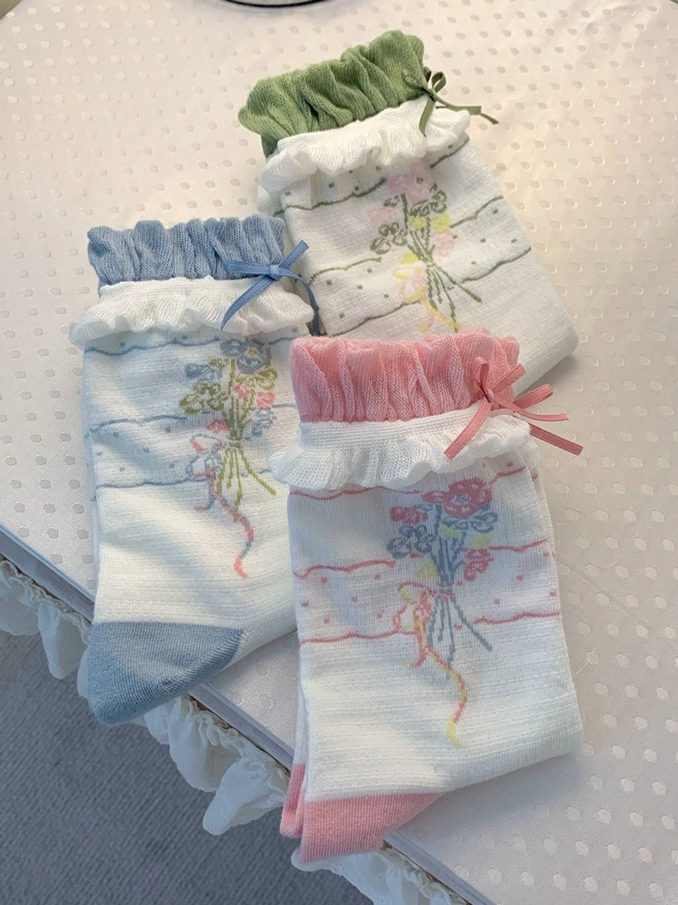 Side flat lay view of three lolita ankle sock variants featuring differently colored ruffled cuffs and heel caps floral bouquet patterns tiny bows on pink cuffs lace-trimmed background