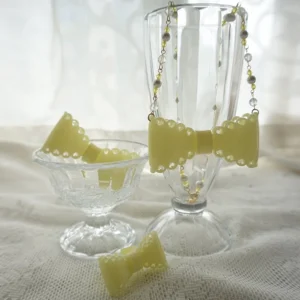 Light yellow matte resin bow necklace with delicate beaded chain and metal accents displayed on vintage glassware and linen tabletop