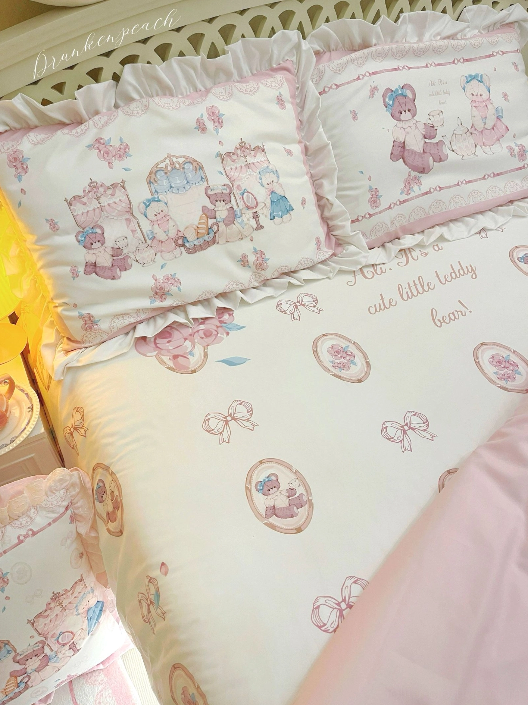Overhead flat-lay display of loose lolita bear bedding sheet with off-white base, pink bow and circular medallion prints, central large bear afternoon tea panel, and neat repeating motif layout