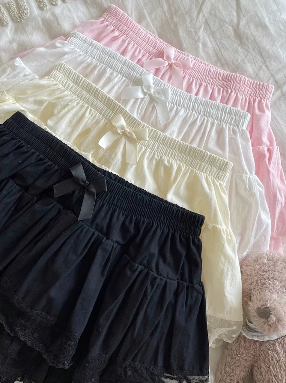 Assorted color Lolita bloomers top-down flat lay display on bedding background with teddy bear decor showing four color options tiered ruffle design elastic waist bow and lace hem details