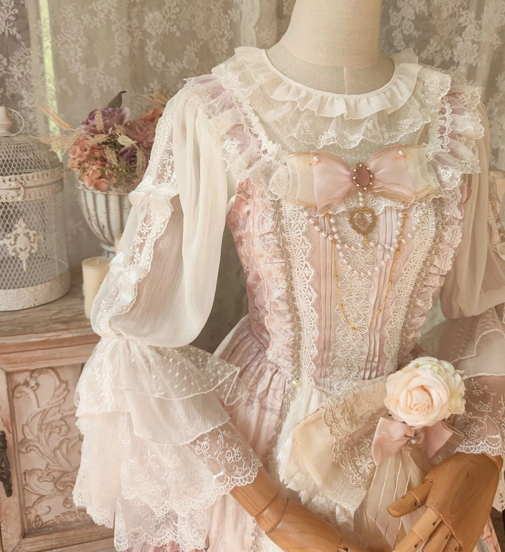 Front close-up three-quarter view of Lolita blouse with pearl and heart charm embellishments pink bow and flower waist decoration and intricate lace bodice against vintage wooden cabinet