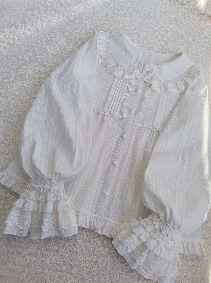 Oblique top-down flat lay of white long-sleeve lolita blouse showcasing peter pan lace collar, front button placket, tiered ruffle lace cuffs, lantern sleeves, and vertical stripe woven cotton fabric on lace background