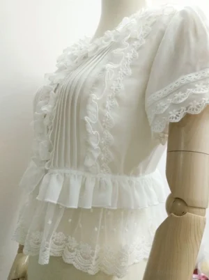 Side profile of romantic ruffled lolita blouse displaying the architectural layered waist ruffle construction, dual-tier hem with dotted tulle mesh overlay creating bell-shaped silhouette, lace-trimmed puffed short sleeve detail, and overall graceful chiffon drape and movement