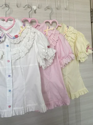Multiple lolita blouses in white pink and pale yellow with lace ruffled collars puffed sleeves front buttons and ruffled hem edges hanging on heart-shaped hangers wardrobe display collection