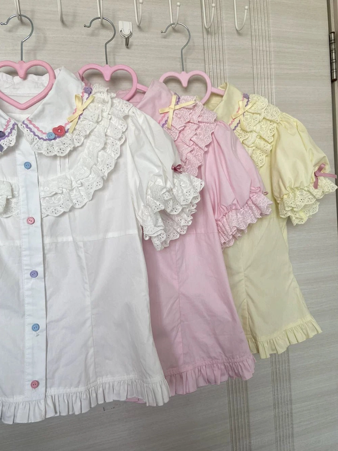 Multiple lolita blouses in white pink and pale yellow with lace ruffled collars puffed sleeves front buttons and ruffled hem edges hanging on heart-shaped hangers wardrobe display collection