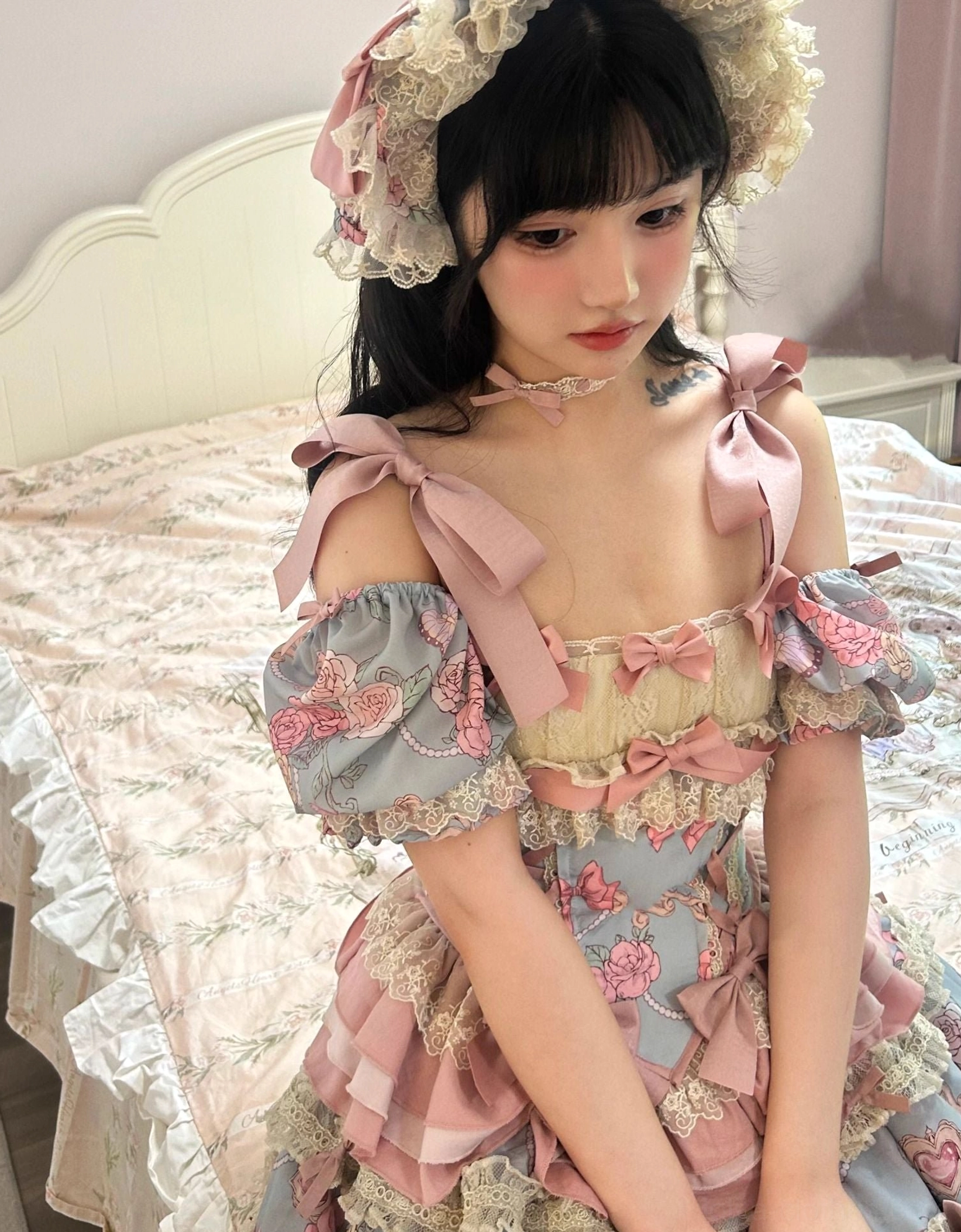 Close-up upper-body shot of sweet lolita dress bodice and lace ruffled bonnet headdress showing square neckline with small pink bows and puff floral print sleeves on bedroom bed background