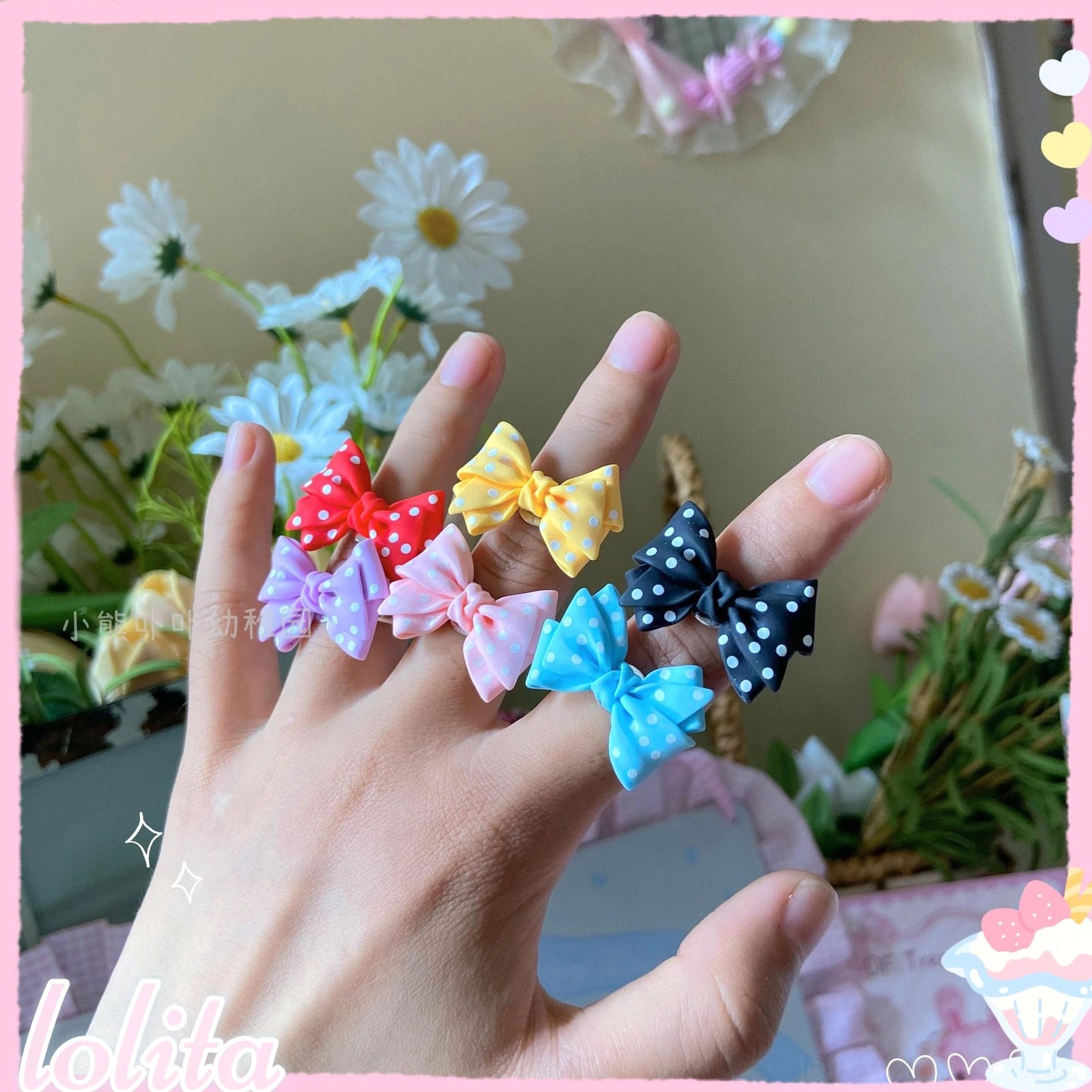 First-person hand-wearing view of five different sweet lolita bowknot statement rings on female fingers with outdoor floral background featuring white daisies and green foliage
