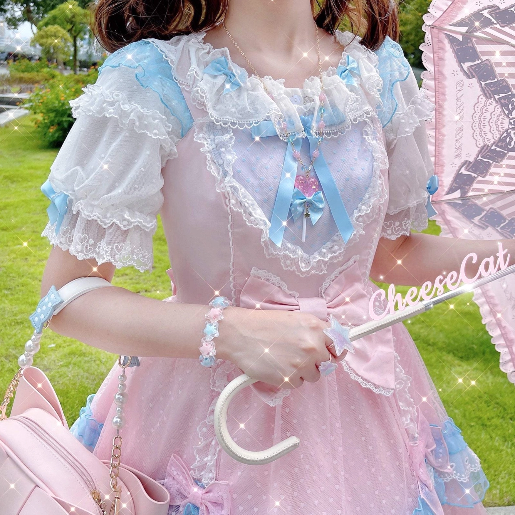 Medium upper-body outdoor shot of girl in pink-blue Lolita dress wearing matching bracelet and accessories with pink bow handbag and lace parasol, showing perfectly coordinated sweet Lolita ensemble