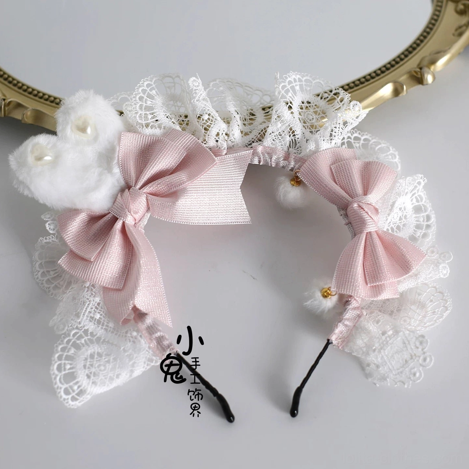 Lolita bunny ear KC hair accessory with white lace frills, pink satin bows, fluffy pom-poms, and tiny bead decorations displayed on vintage gold mirror