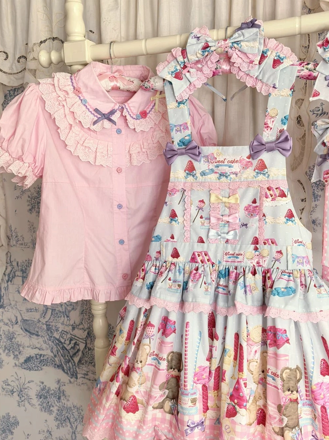 Sweet lolita coordination featuring pink blouse with lace ruffled details paired with sweet printed overall dress with cartoon dessert strawberry and bear prints and purple bow accents